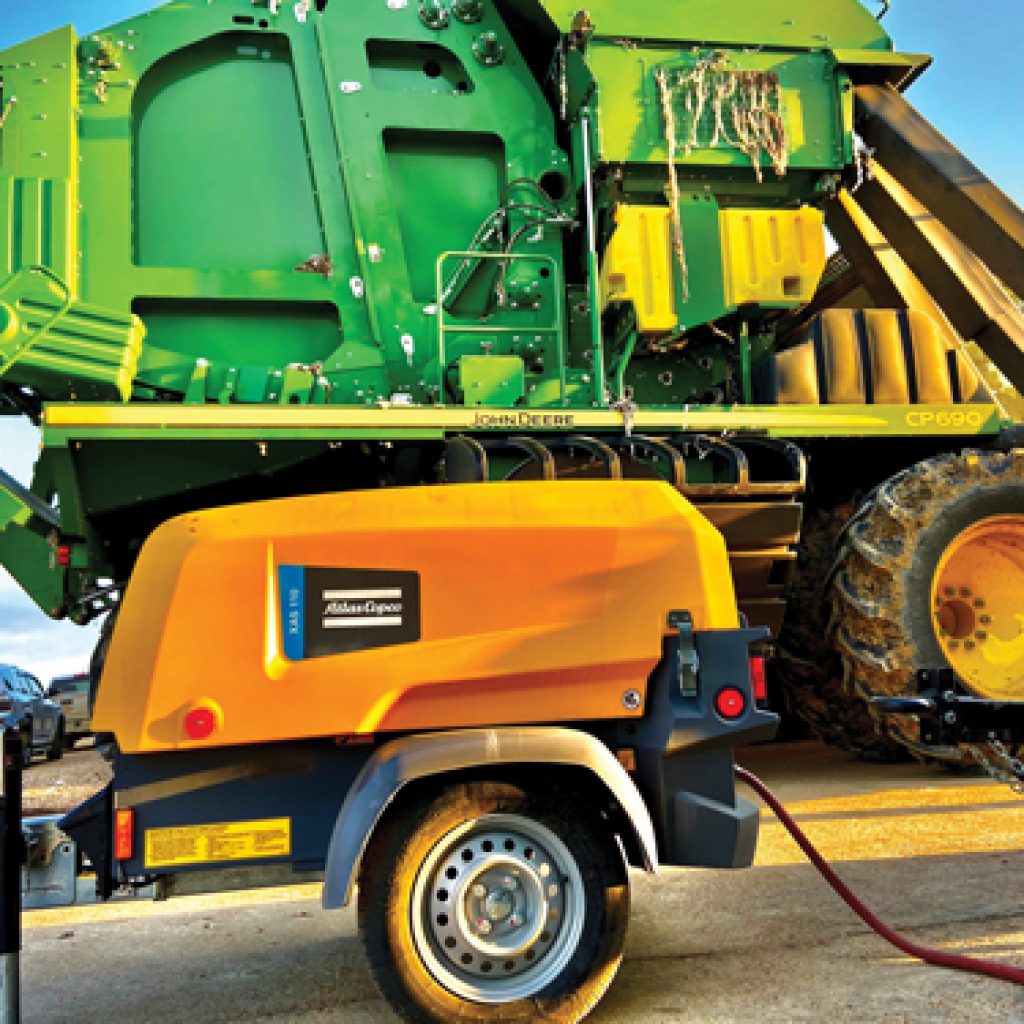 A large green machine with a trailer attached, set against a clear sky and grassy area.