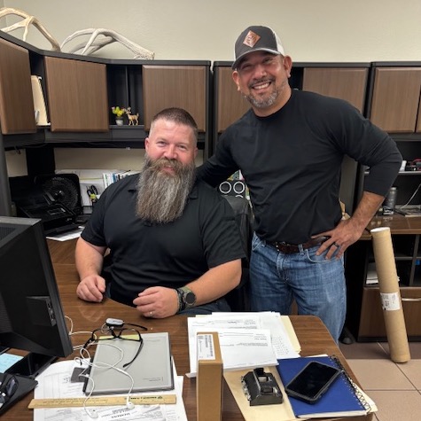 Two men working in an office, focused on their computers surrounded by papers and office supplies.